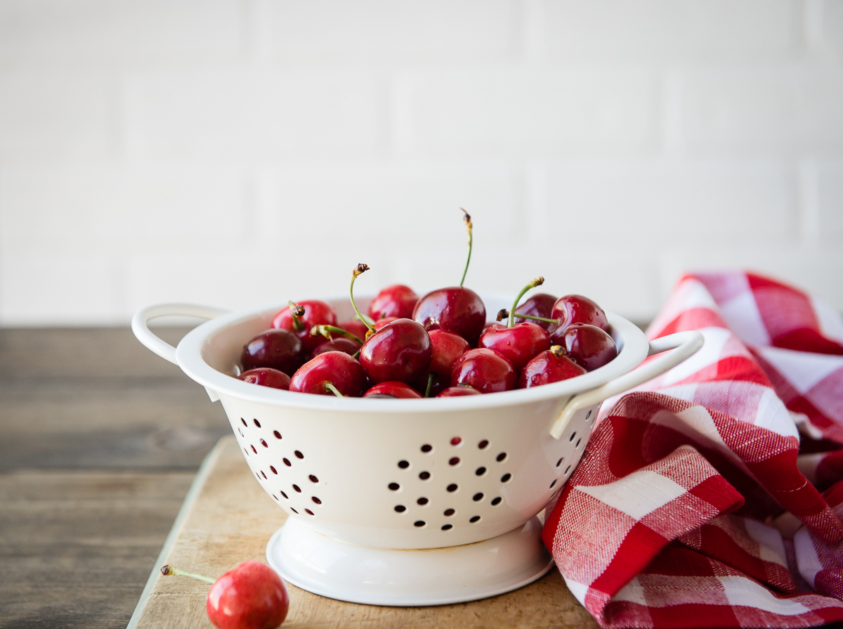 white enamel strainer full of red cherries, on a wooden cutting board, with a red and white checked towel